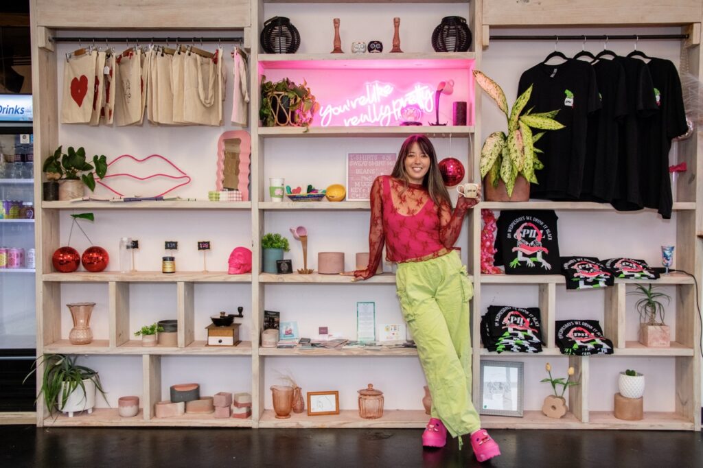 Woman in chartruese pants and a pink top stands in front of a book case holding a cup of coffee