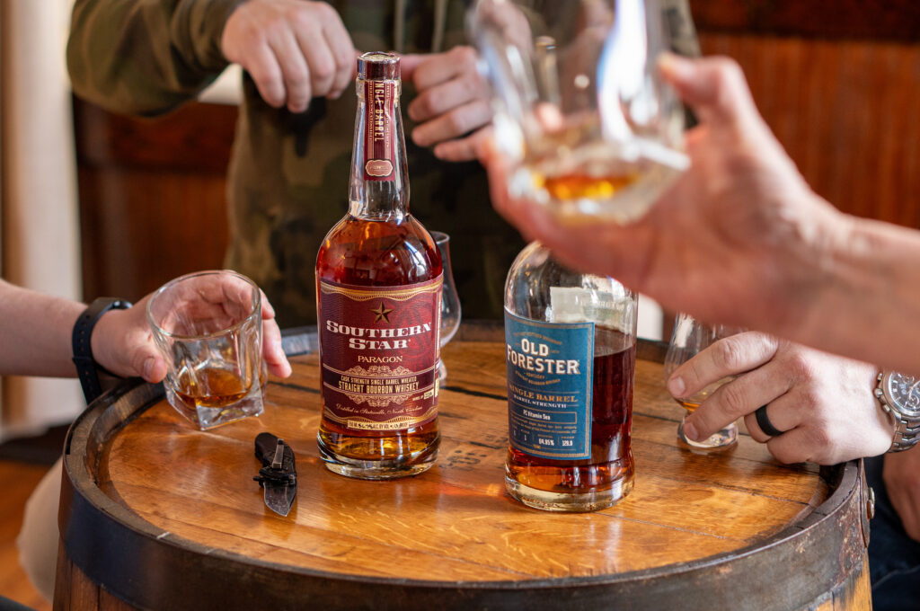 top bottles of whiskey sit on top of a barrel, three hands hold glasses to cheers